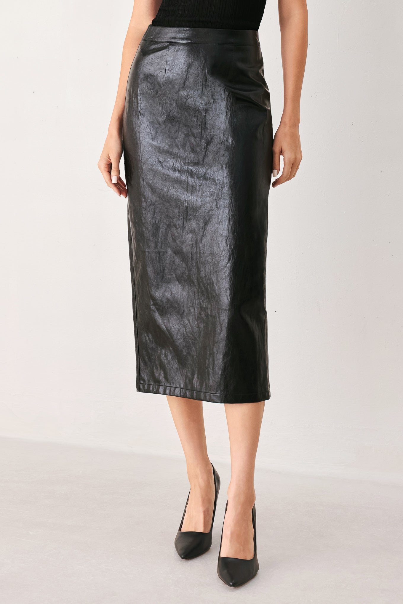 A black faux-leather midi skirt worn by a model, paired with black pointed-toe heels, shown against a plain light gray studio background.