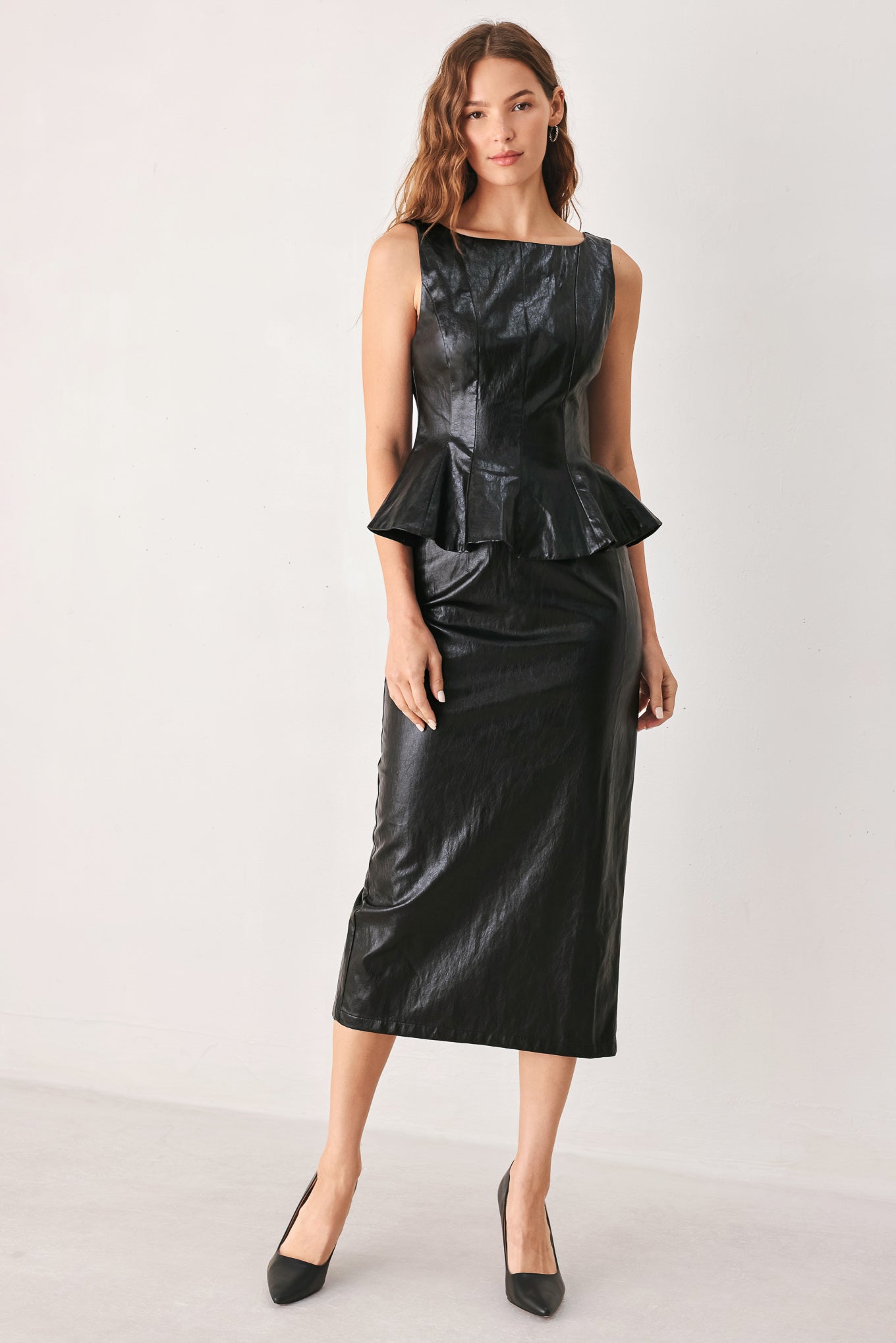 A woman modeling a sleeveless black faux-leather midi dress with a structured bodice, peplum waist, and straight skirt, paired with black pointed-toe heels against a plain light gray studio background.