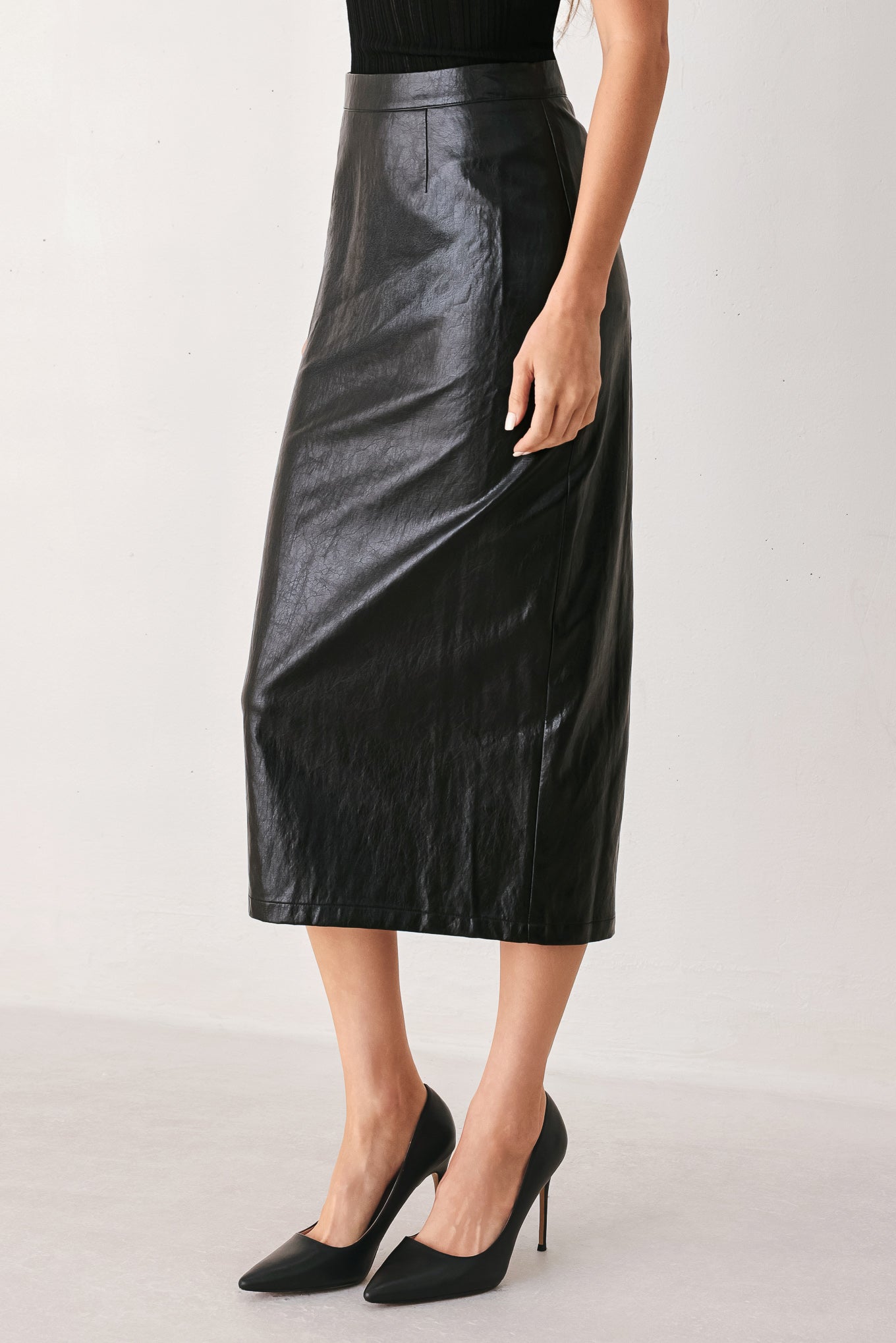 A model wearing a black faux-leather midi skirt with a subtle sheen, paired with black pointed-toe heels, shown from the waist down against a light gray studio background.