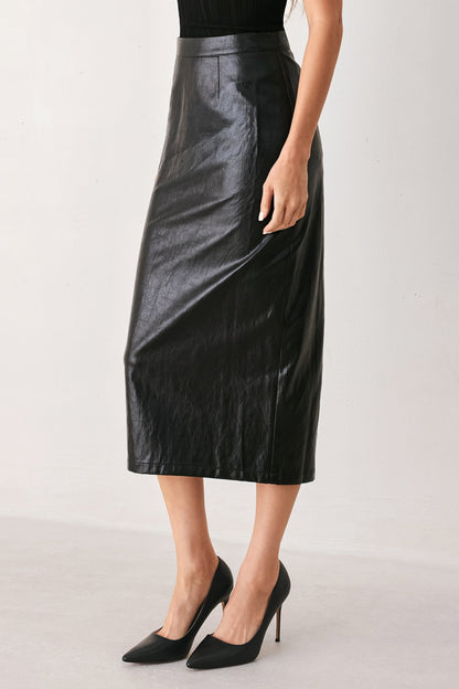 A model wearing a black faux-leather midi skirt with a subtle sheen, paired with black pointed-toe heels, shown from the waist down against a light gray studio background.