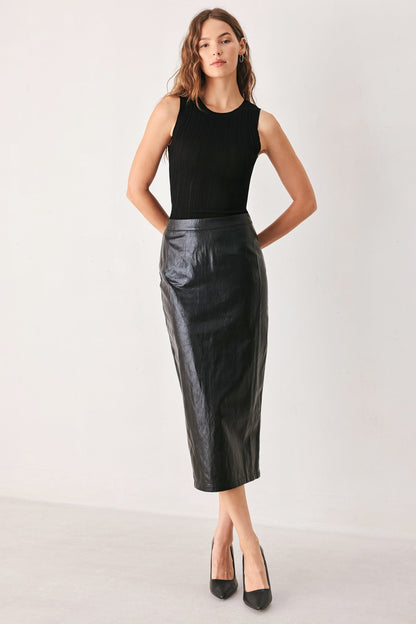 A model wearing a black faux-leather midi skirt with a subtle sheen, paired with black pointed-toe heels, shown from the waist down against a light gray studio background.