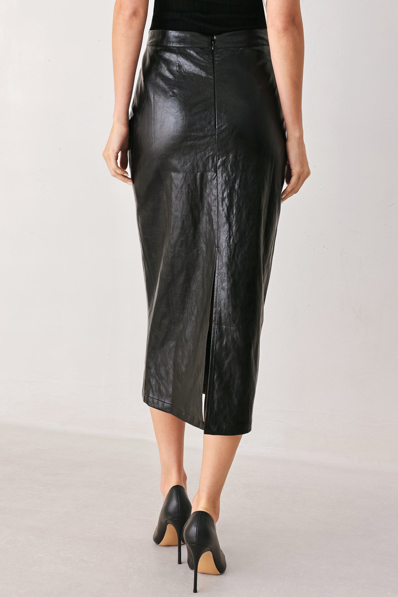 A model from the back wearing a black faux-leather midi skirt with a front zipper and side slit, paired with black pointed-toe heels, against a light gray studio background.