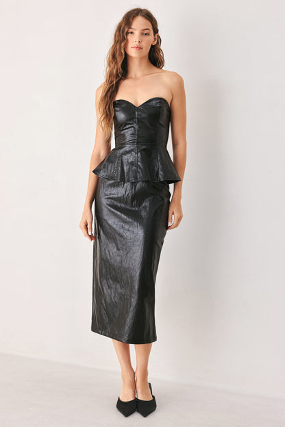 A woman modeling a black strapless faux-leather midi dress with a sweetheart neckline, structured corset-style bodice, peplum waist, and fitted skirt, paired with black pointed-toe heels against a plain light gray studio background.