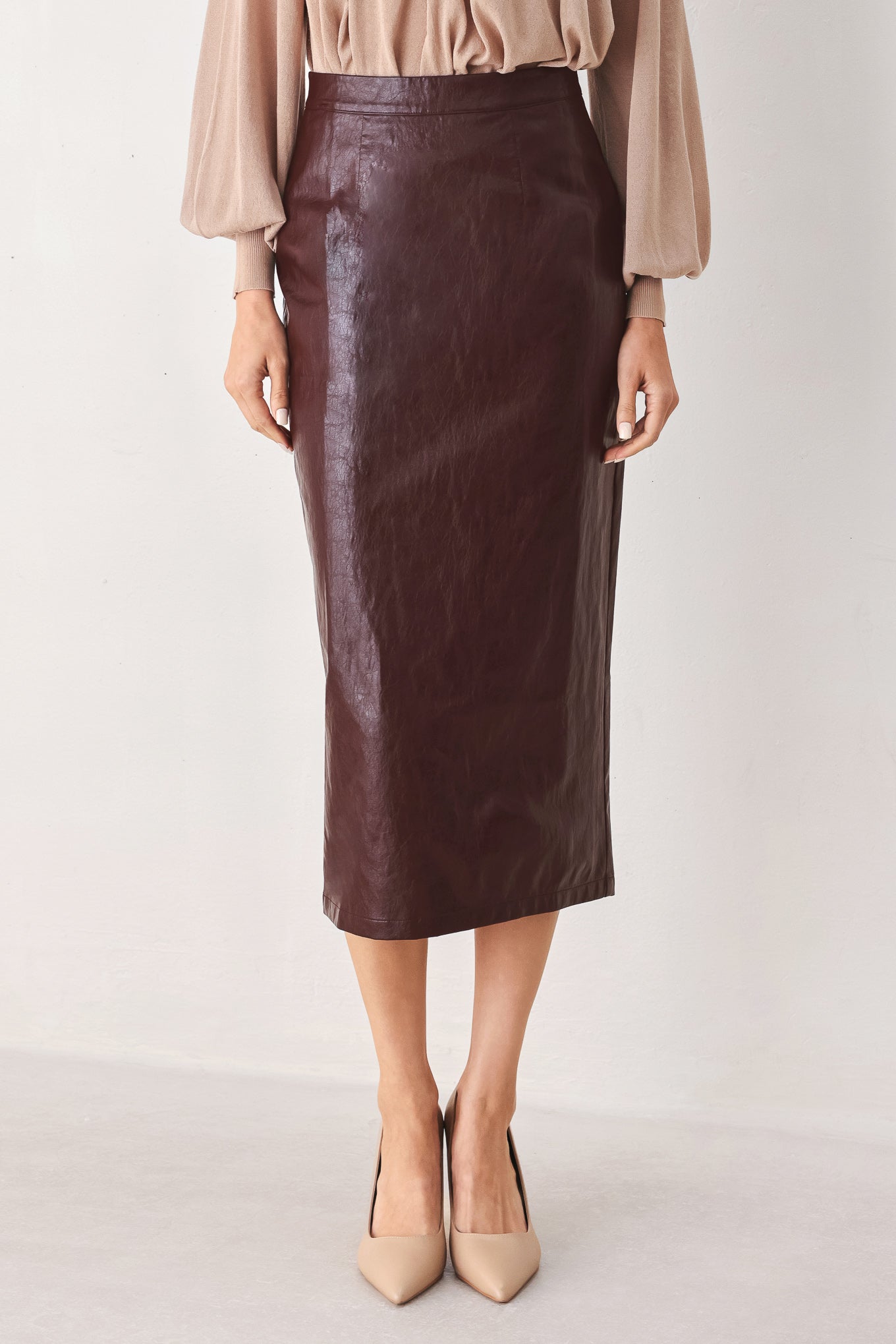 A model wearing a dark brown faux-leather midi skirt paired with a beige blouse and beige pointed-toe heels, shown from the waist down against a plain off-white studio background.