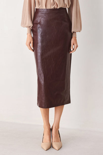 A model wearing a dark brown faux-leather midi skirt paired with a beige blouse and beige pointed-toe heels, shown from the waist down against a plain off-white studio background.