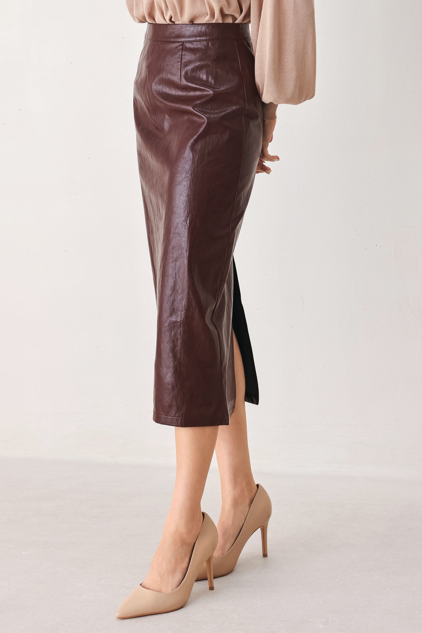 A model wearing a dark brown faux-leather midi skirt with a side slit, paired with beige pointed-toe heels and a beige top, shown from the waist down against a plain light background.
