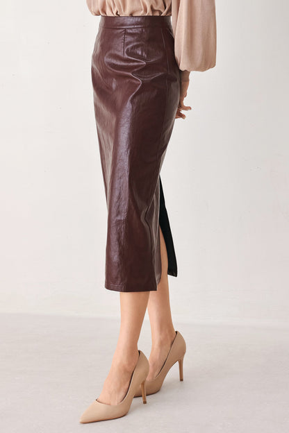 A model wearing a dark brown faux-leather midi skirt with a side slit, paired with beige pointed-toe heels and a beige top, shown from the waist down against a plain light background.