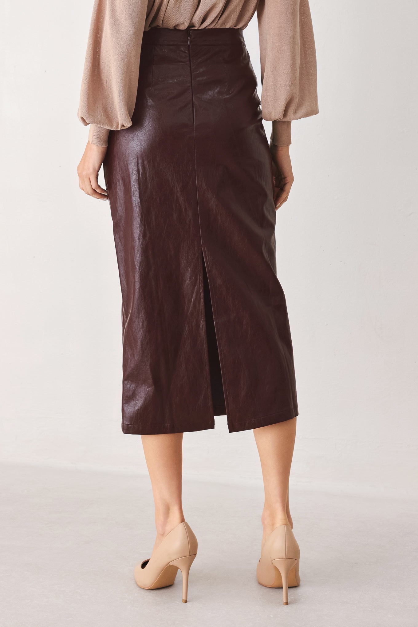 A model from the back wearing a dark brown faux-leather midi skirt with a front slit, paired with a beige blouse and beige heels, against a light gray studio background.