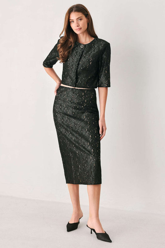 A woman models a black two-piece lace suit featuring a short-sleeve cropped top with a buttoned round neckline and a matching knee-length pencil skirt, styled with a thin metallic belt and black pointed-toe mule heels against a light gray studio background.