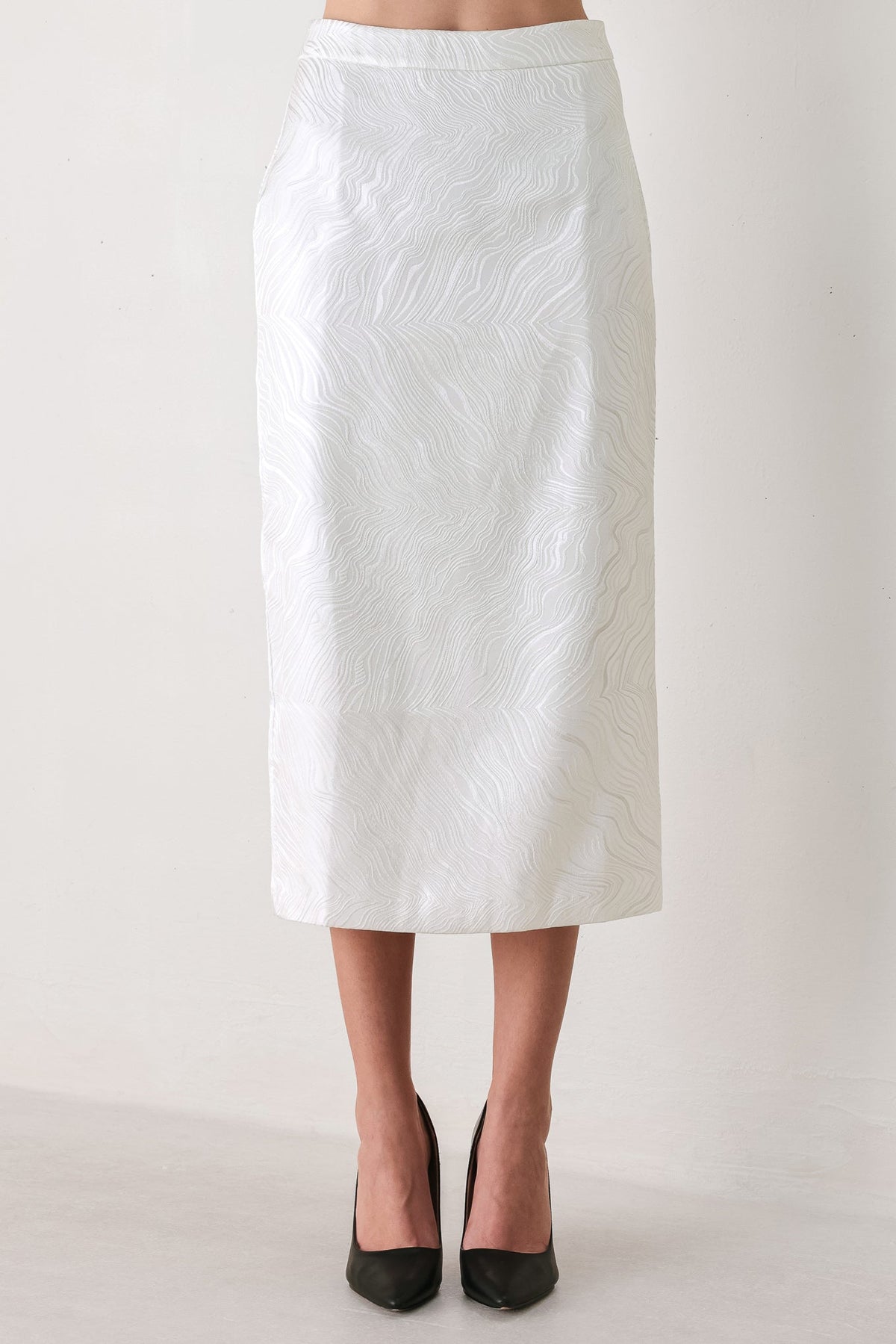 A white midi skirt with a subtle textured, wavy jacquard pattern, shown on a model wearing black pointed-toe heels against a plain off-white studio background.