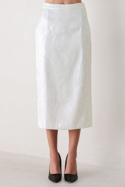 A white midi skirt with a subtle textured, wavy jacquard pattern, shown on a model wearing black pointed-toe heels against a plain off-white studio background.
