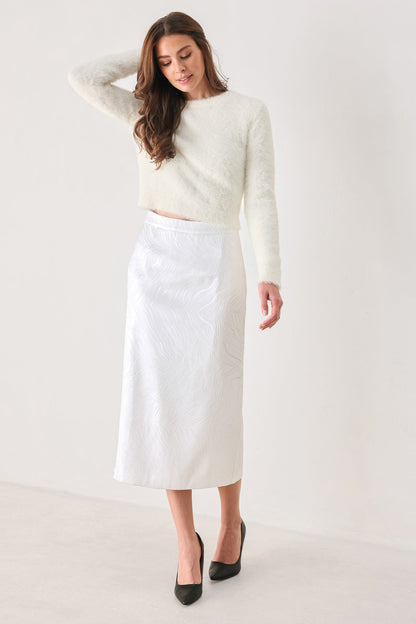 A woman modeling a white textured long-sleeve crop top and a white textured midi skirt, paired with black pointed-toe heels, against a plain off-white studio background.