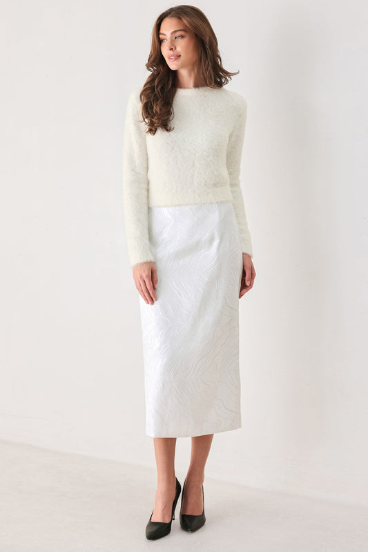A woman modeling a cream-colored textured knit sweater paired with a white textured midi skirt and black pointed-toe heels, set against a plain off-white studio background.