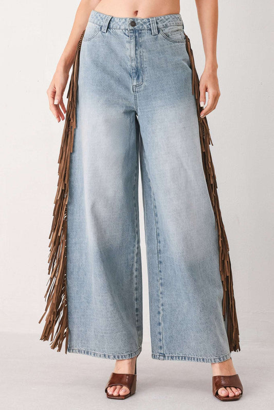 A model wearing light-wash blue wide-leg denim jeans with long brown fringe detailing running down the outer sides, paired with brown heeled sandals against a plain light gray studio background.