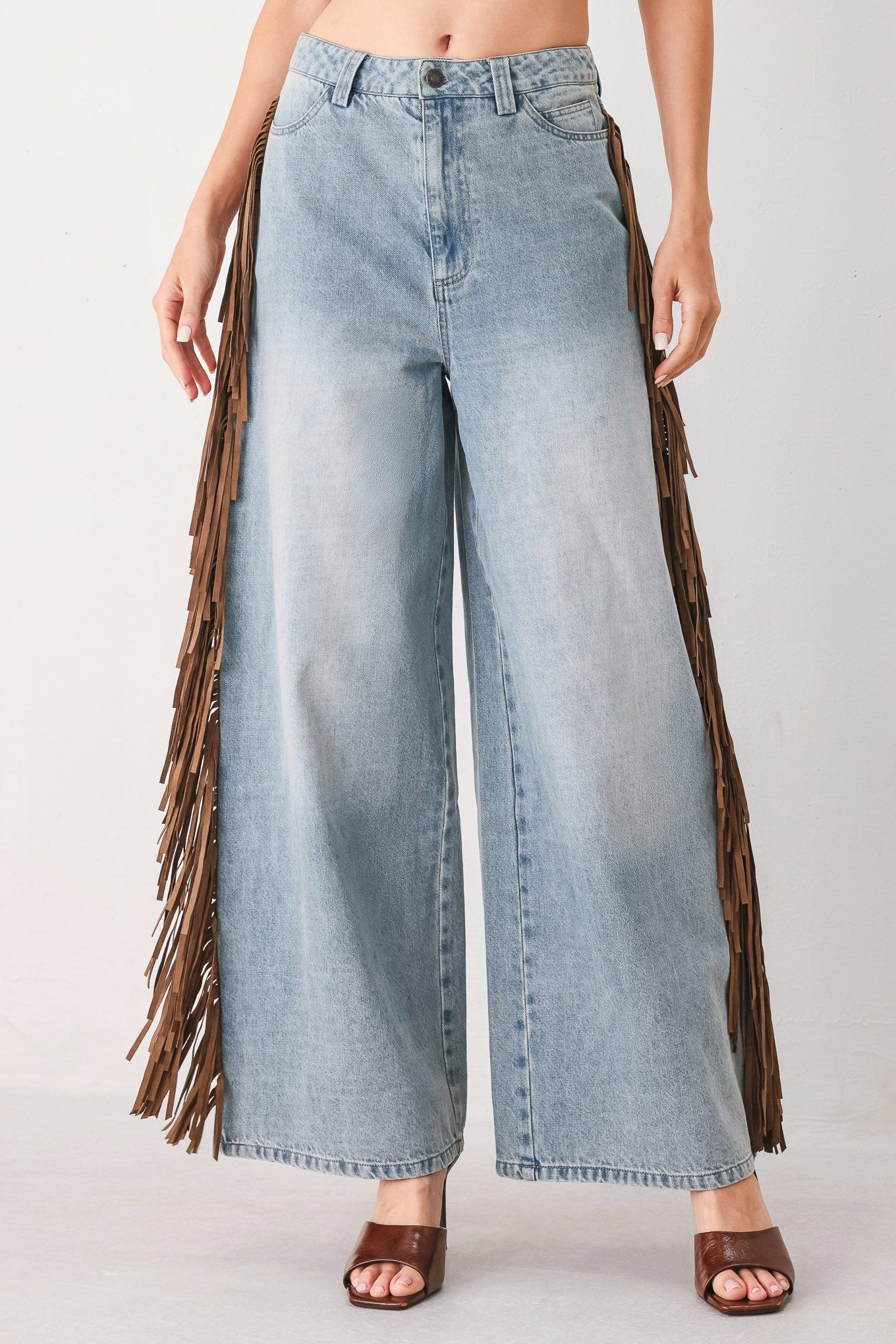 A model wearing light-wash blue wide-leg denim jeans with long brown fringe detailing running down the outer sides, paired with brown heeled sandals against a plain light gray studio background.