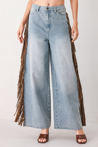 A model wearing light-wash blue wide-leg denim jeans with long brown fringe detailing running down the outer sides, paired with brown heeled sandals against a plain light gray studio background.