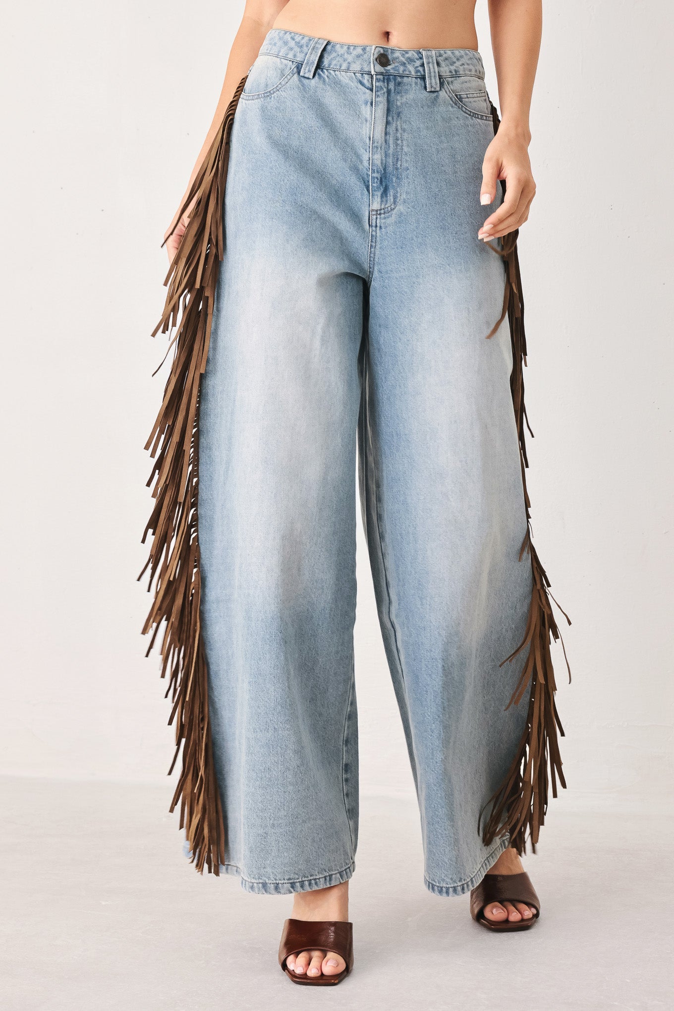 A model wearing light-wash wide-leg denim jeans with long brown fringe hanging down the outer sides, paired with brown slide sandals against a plain light gray studio background.