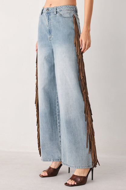 Light-wash wide-leg denim jeans with long brown fringe detailing running down the outer sides, shown on a model wearing brown heeled sandals against a plain light gray studio background.