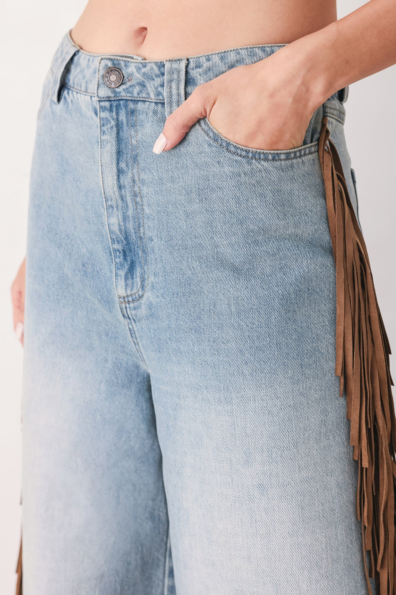 Close-up of a person wearing light-wash blue denim jeans with a brown fringe detail running down the side, with one hand in the pocket.