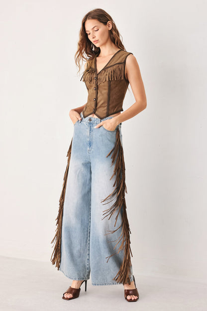 A woman modeling a brown fringed sleeveless top and matching wide-leg light blue denim jeans with long brown fringe, paired with brown heeled sandals, against a plain light gray studio background.