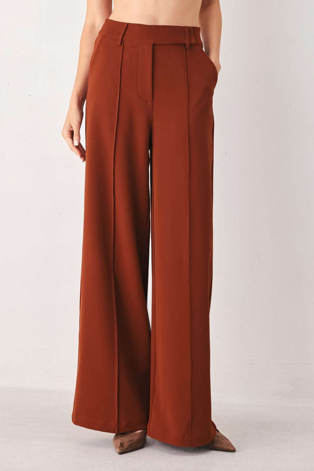 Rust-colored wide-leg fabric trousers with front seams and belt loops worn against a white wall.