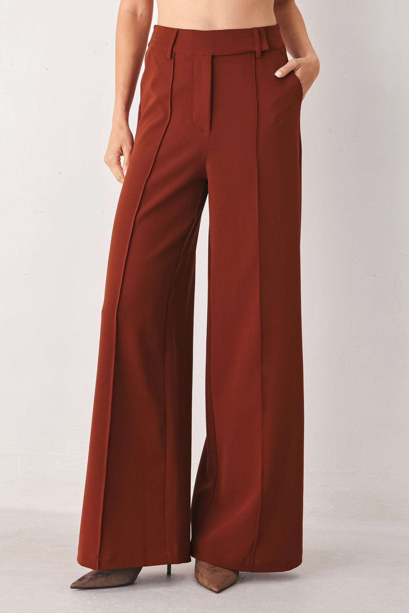 Rust brown wide-leg fabric pants with front seams, belt loops, and side pockets worn against a white wall.