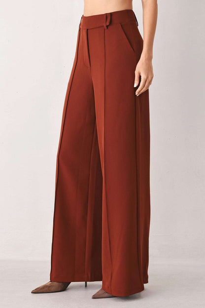 Rust brown fabric wide-leg trousers with front pleats and belt loops, worn with pointed-toe heels.