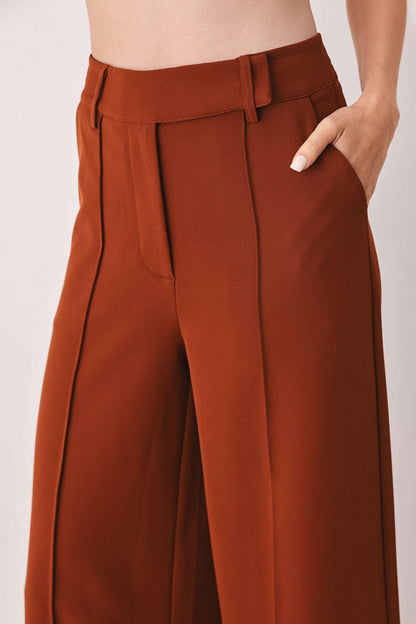 Close-up of rust brown fabric trousers with a high waist, front seams, and a hand in the side pocket.