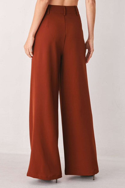 Rear view of a person wearing rust-colored, wide-leg fabric pants with belt loops against a white wall.