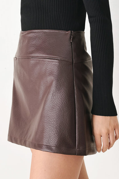 A close-up of a model wearing a brown faux-leather mini skirt with a pebbled texture, paired with a black ribbed long-sleeve top, against a plain white background.