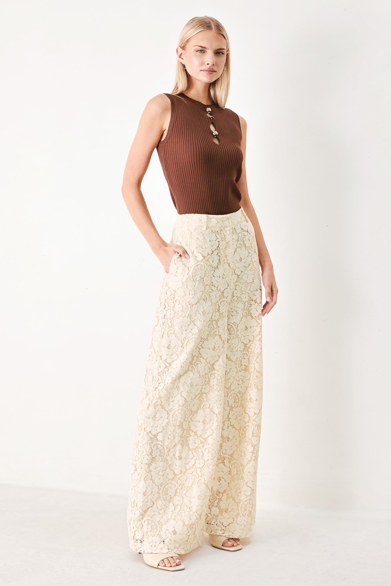 Woman wearing a brown sleeveless top and cream lace pants on a white background