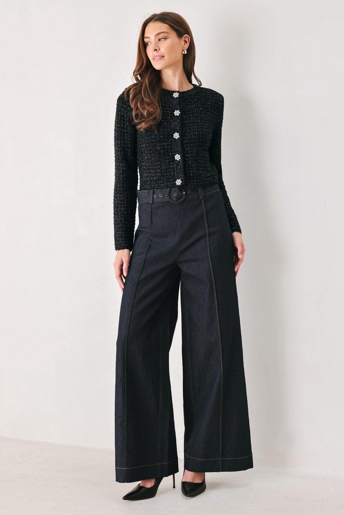 A woman models a black textured knit long-sleeve top with silver buttons and black wide-leg trousers with contrast stitching, standing against a plain light gray studio background.