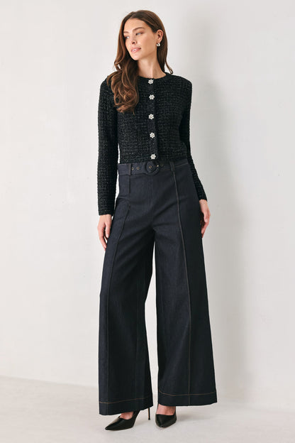 A woman models a black textured knit long-sleeve top with silver buttons and black wide-leg trousers with contrast stitching, standing against a plain light gray studio background.