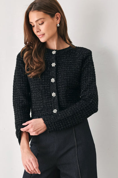 A woman wearing a black tweed cropped jacket with decorative silver buttons down the front, paired with dark trousers, photographed against a plain light gray wall.
