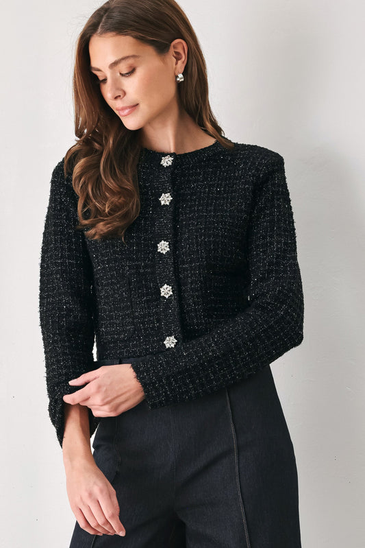 A woman wearing a black tweed cropped jacket with decorative silver buttons down the front, paired with dark trousers, photographed against a plain light gray wall.