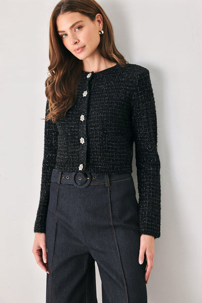 A woman wearing a black textured tweed cardigan with decorative silver buttons and dark charcoal wide-leg trousers, standing against a plain light gray wall.