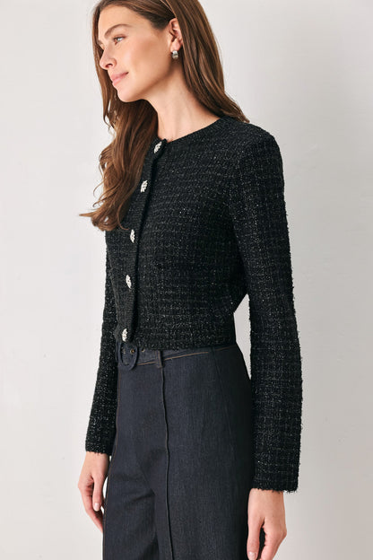 A woman wearing a black textured tweed cardigan with silver buttons and dark charcoal trousers, photographed against a neutral light gray studio background.