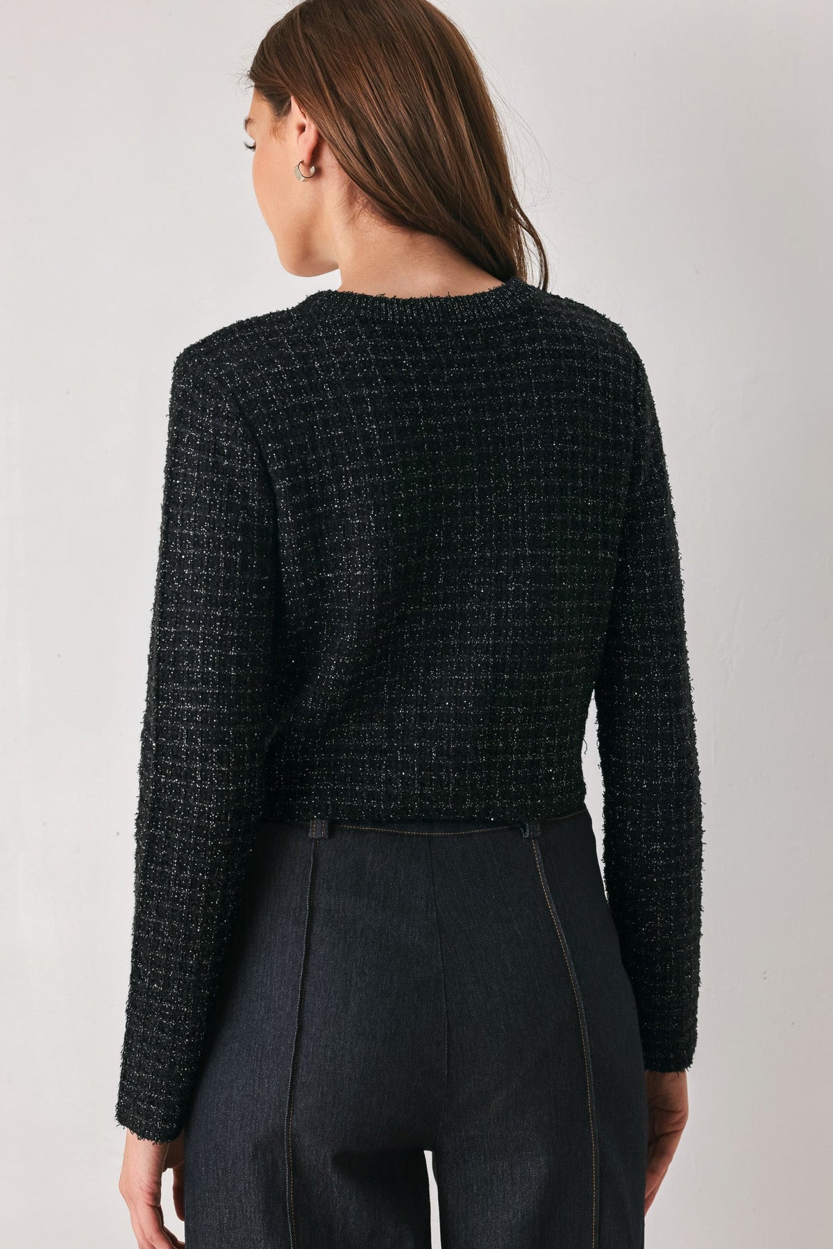 A woman shown from the back wearing a black textured tweed long-sleeve cropped sweater and dark charcoal trousers, photographed against a neutral light gray studio background.