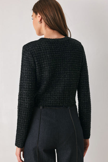 A woman shown from the back wearing a black textured tweed long-sleeve cropped sweater and dark charcoal trousers, photographed against a neutral light gray studio background.