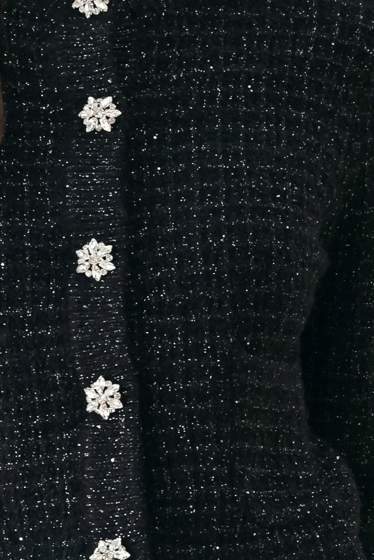 Close-up of a black glittery knit sweater featuring a vertical row of decorative snowflake-shaped buttons embellished with clear rhinestones.