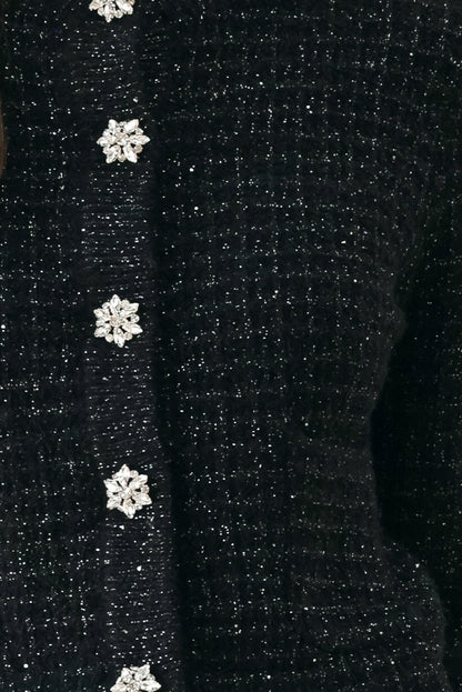 Close-up of a black glittery knit sweater featuring a vertical row of decorative snowflake-shaped buttons embellished with clear rhinestones.