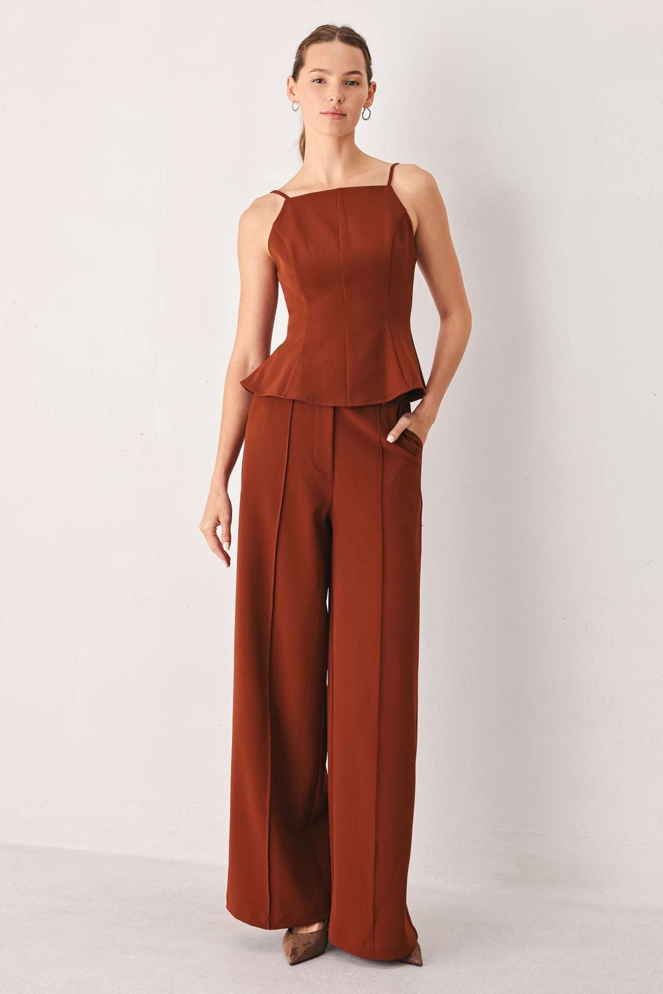 A woman modeling a rust-brown sleeveless jumpsuit with thin shoulder straps, a peplum waist, and wide-leg trousers, standing against a plain light gray studio background.