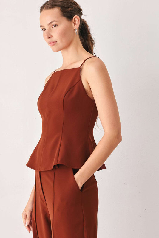 A woman modeling a rust-brown sleeveless peplum top and matching trousers against a plain light gray studio background.