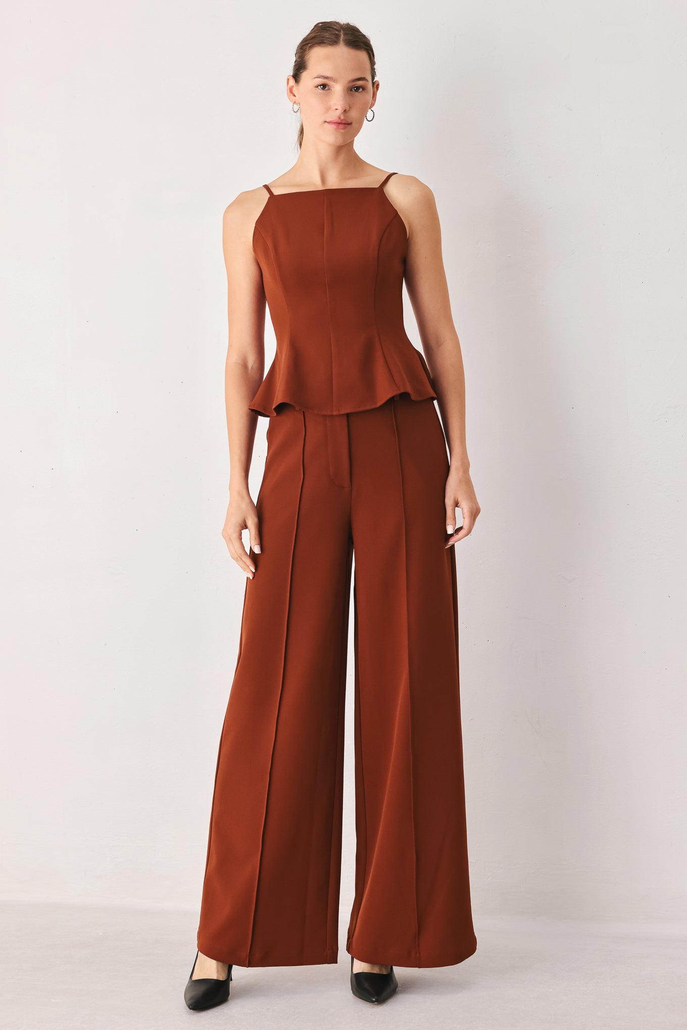 A woman modeling a rust-brown sleeveless jumpsuit with a peplum waist and wide-leg pants against a plain light gray studio background.