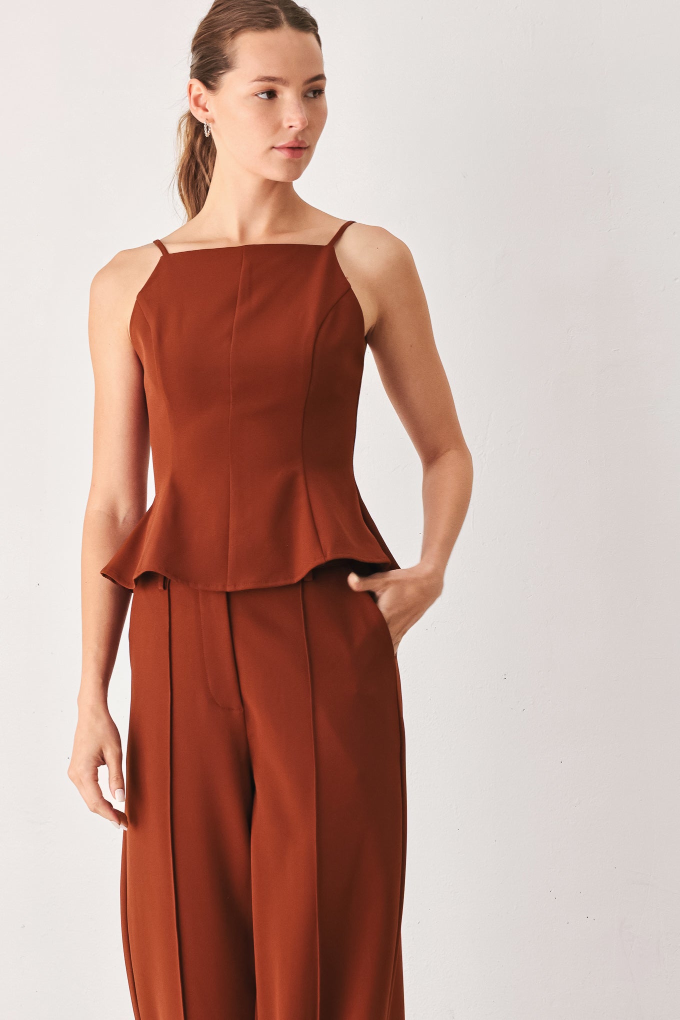 A woman modeling a rust-brown sleeveless peplum top and matching wide-leg pants against a plain light gray studio background.