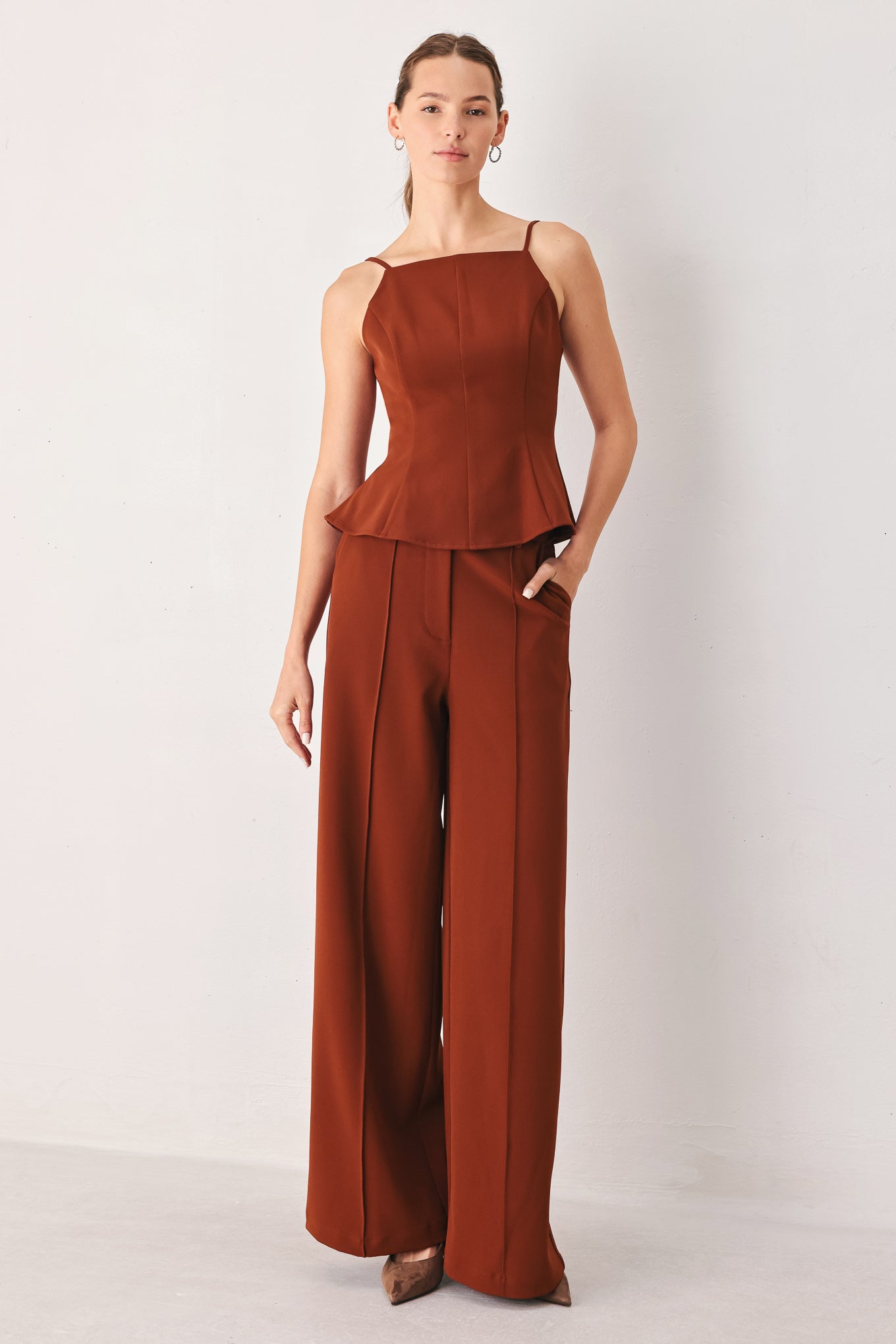 A woman modeling a rust-brown sleeveless jumpsuit with thin shoulder straps, a peplum waist, and wide-leg trousers, standing against a plain light gray studio background.