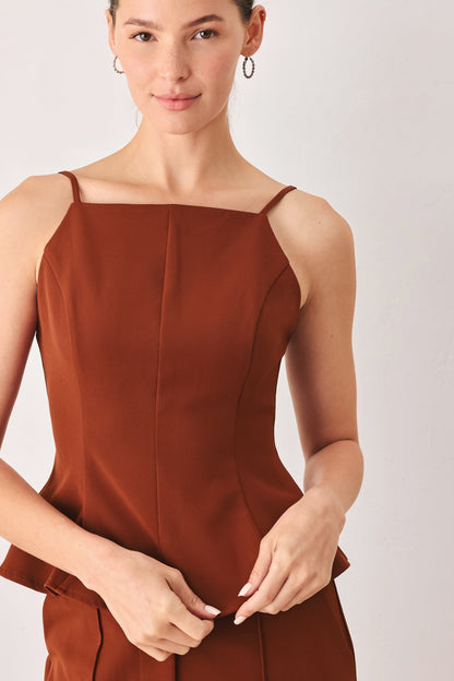 A woman wearing a rust-brown sleeveless camisole-style top with thin straps and a structured square neckline, photographed against a light neutral studio background.