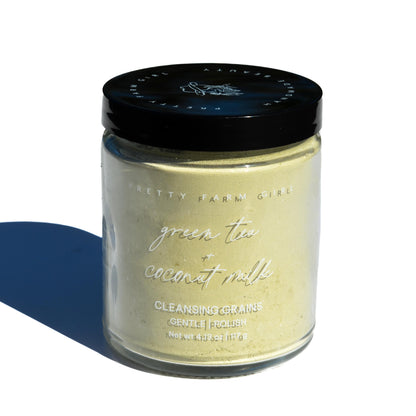 Jar of 'Green Tea + Coconut Milk' cleansing granules with a black lid on a white background
