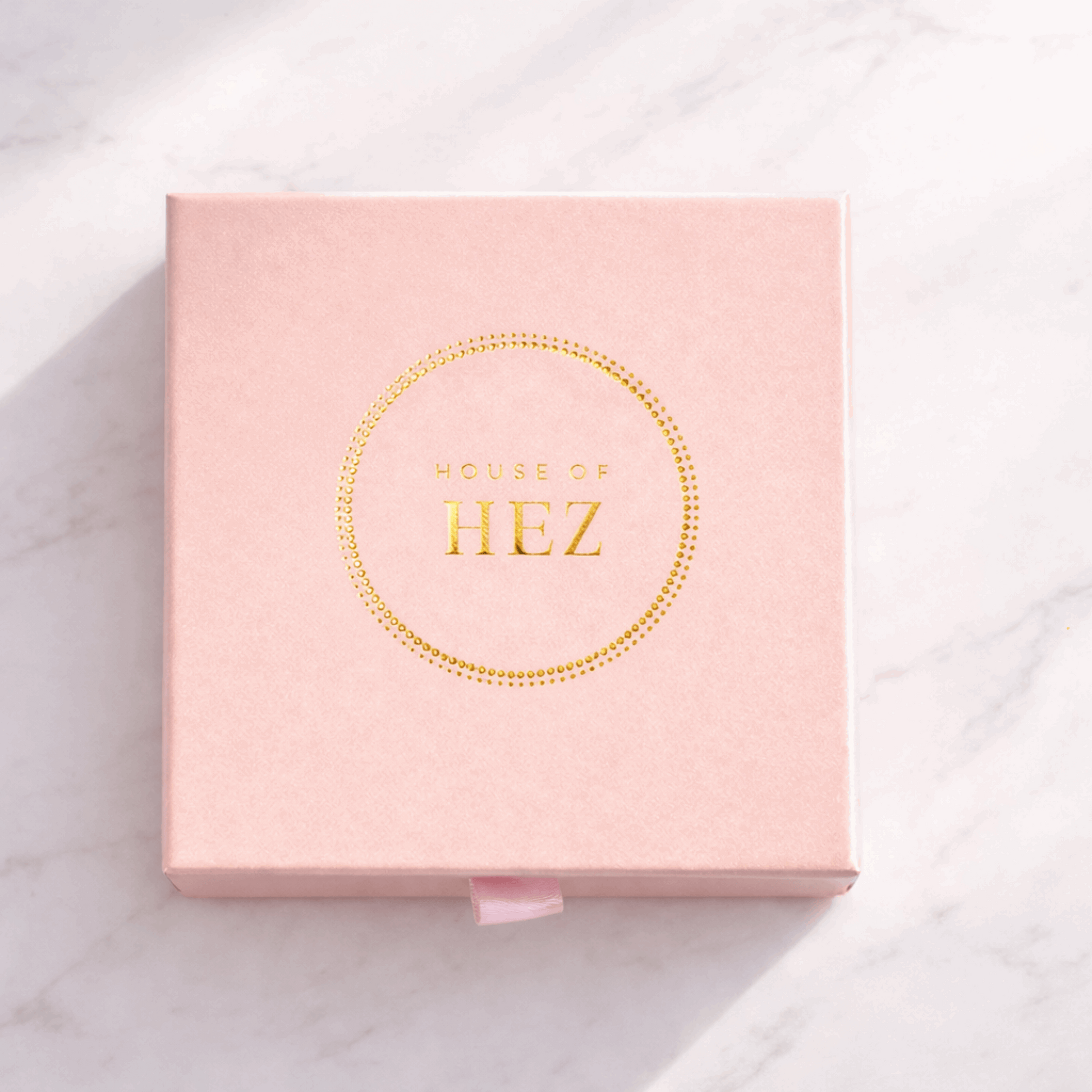 Square pink box with a gold foil HOUSE OF HEZ logo inside a dotted circle, resting on a white marble surface.