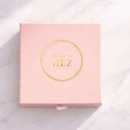 Square pink box with a gold foil HOUSE OF HEZ logo inside a dotted circle, resting on a white marble surface.